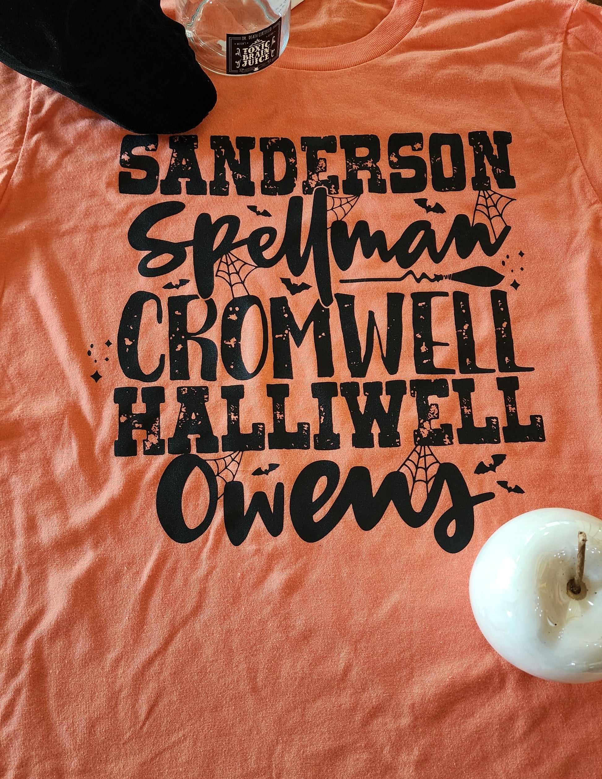 Pictured: Tee is orange with black writing that reads, Sanderson, Spellman, Cromwell, Halliwell, and Owens. 