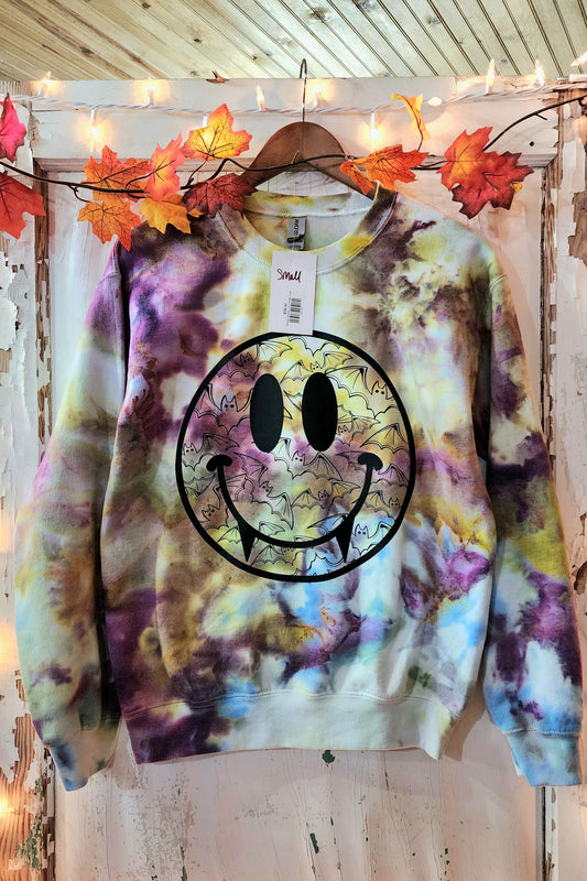 Tie dye sweat shirt in colors of brown, blue, purple, and hints of green. Large smiley face is pressed to the front with vampire teeth. Bat designs are contain in the smiley