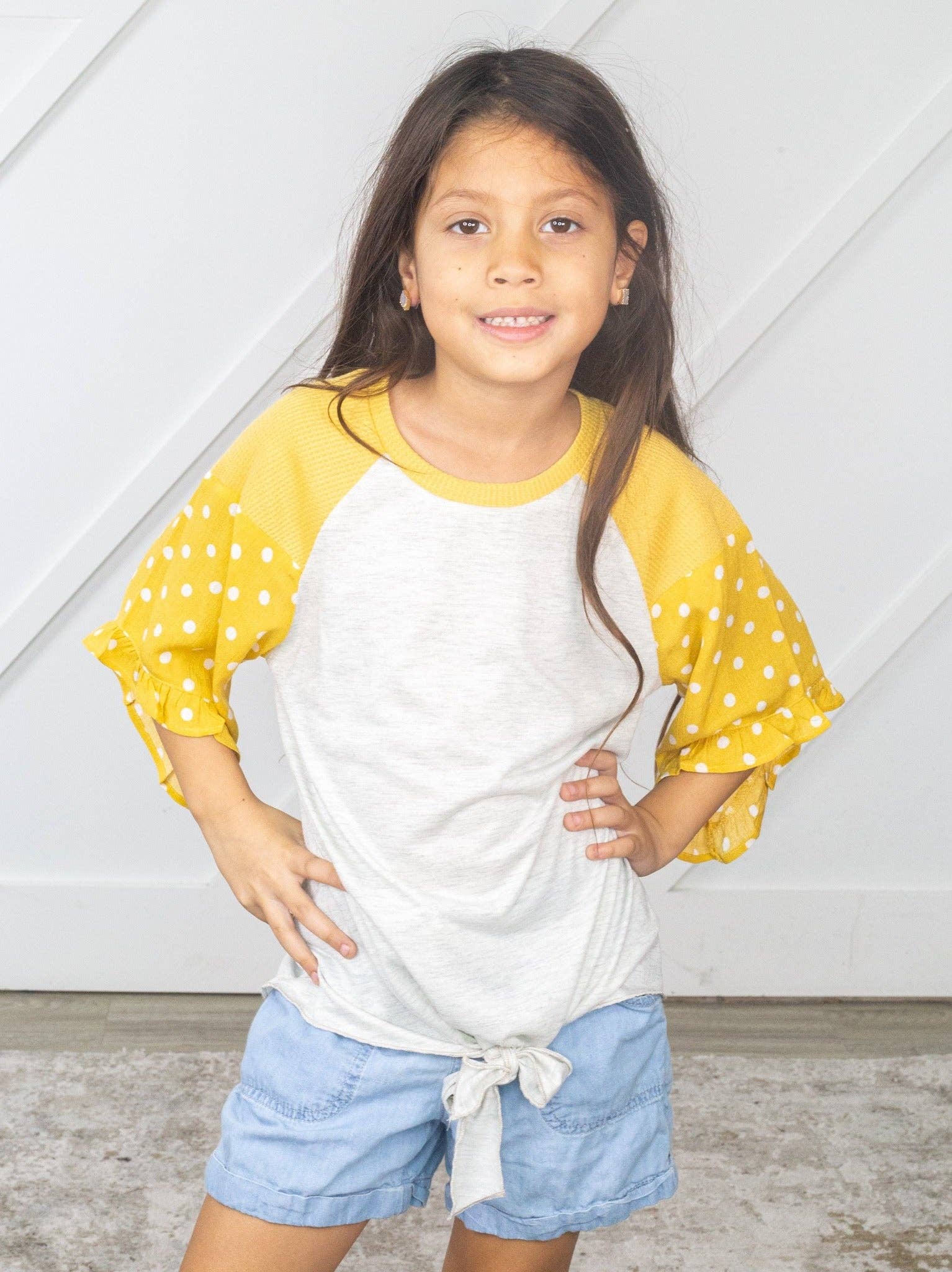 Girl modeling grey and yellow shirt. White polka dots on sleeves along with ruffles. Shirt tied at the bottom. 